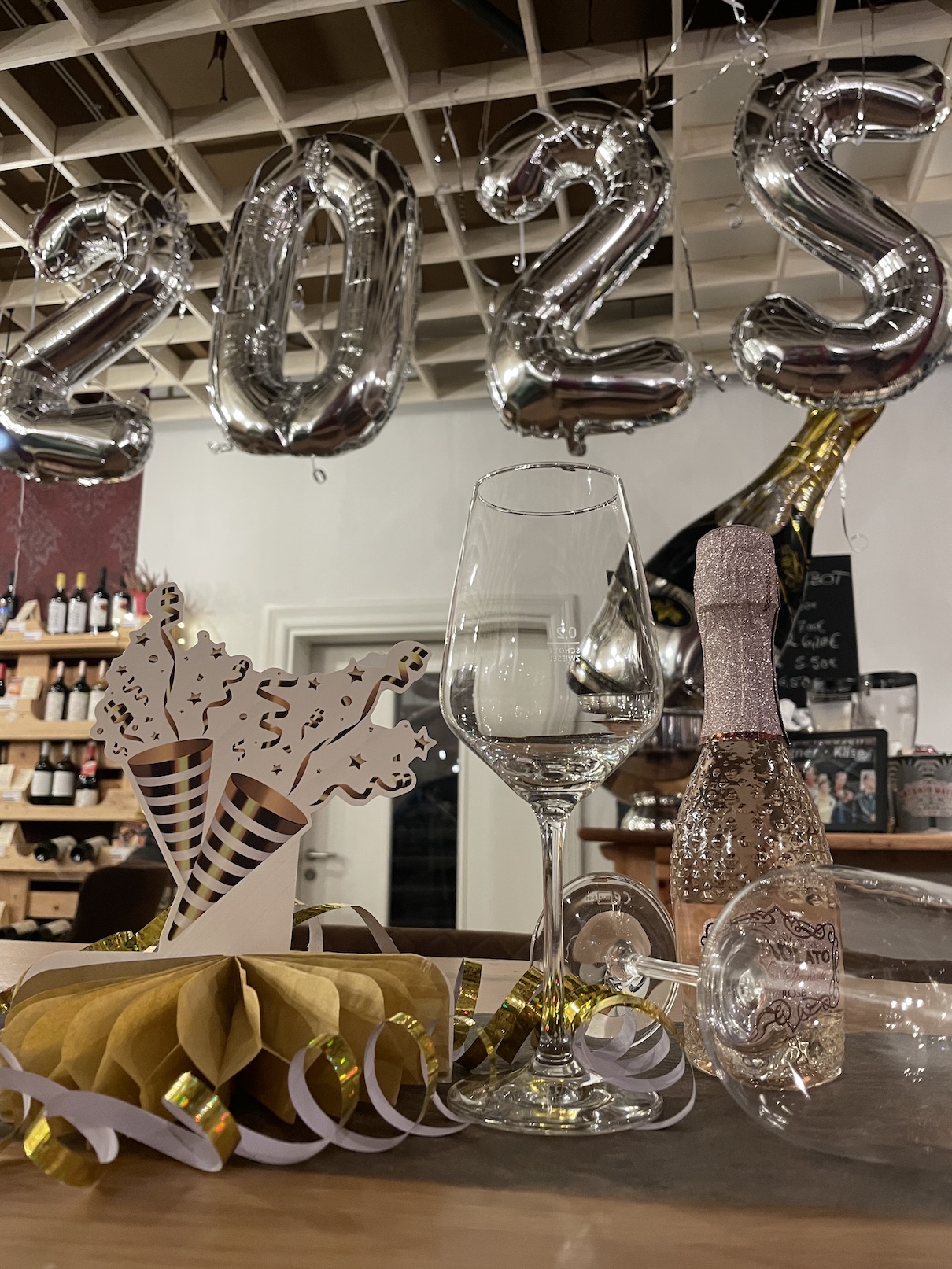 Zwei Weingläser und Silvester-Dekoration liegen auf einem Tisch. Darüber ist mit Luftballons 2025 geschrieben.
